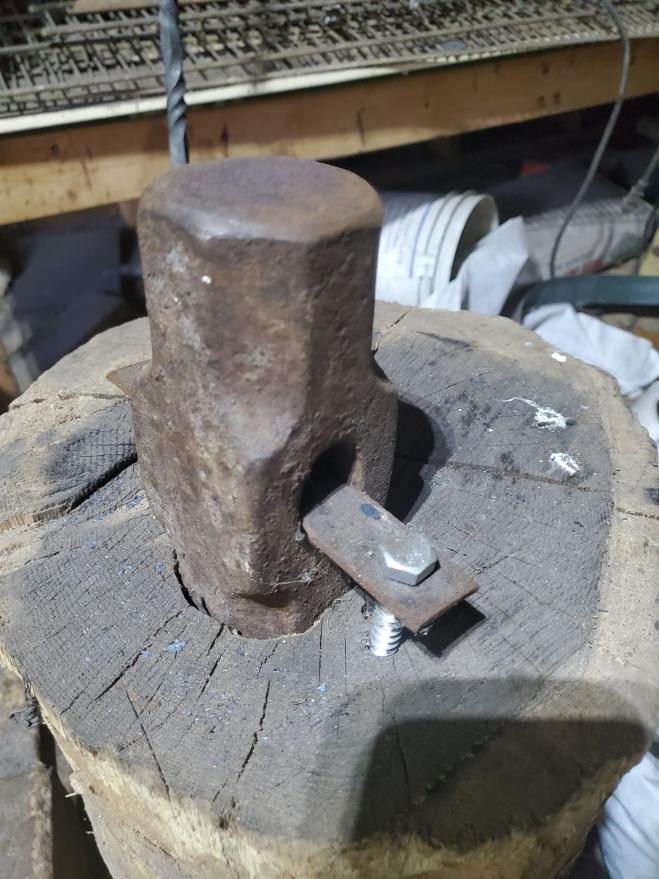 A sledgehammer head sits upright in a hole drilled in the end of a log. The sledgehammer head is affixed to the log by means of a flat, rectangular strip of metal threaded through the eye, which is held to the log by two lag screws.