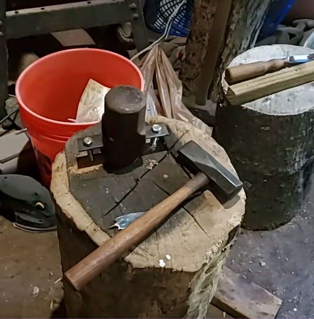 An oblique, zoomed out view of the mounted sledgehammer anvil. A diagonal crosspein hammer sits on the stump next to it.