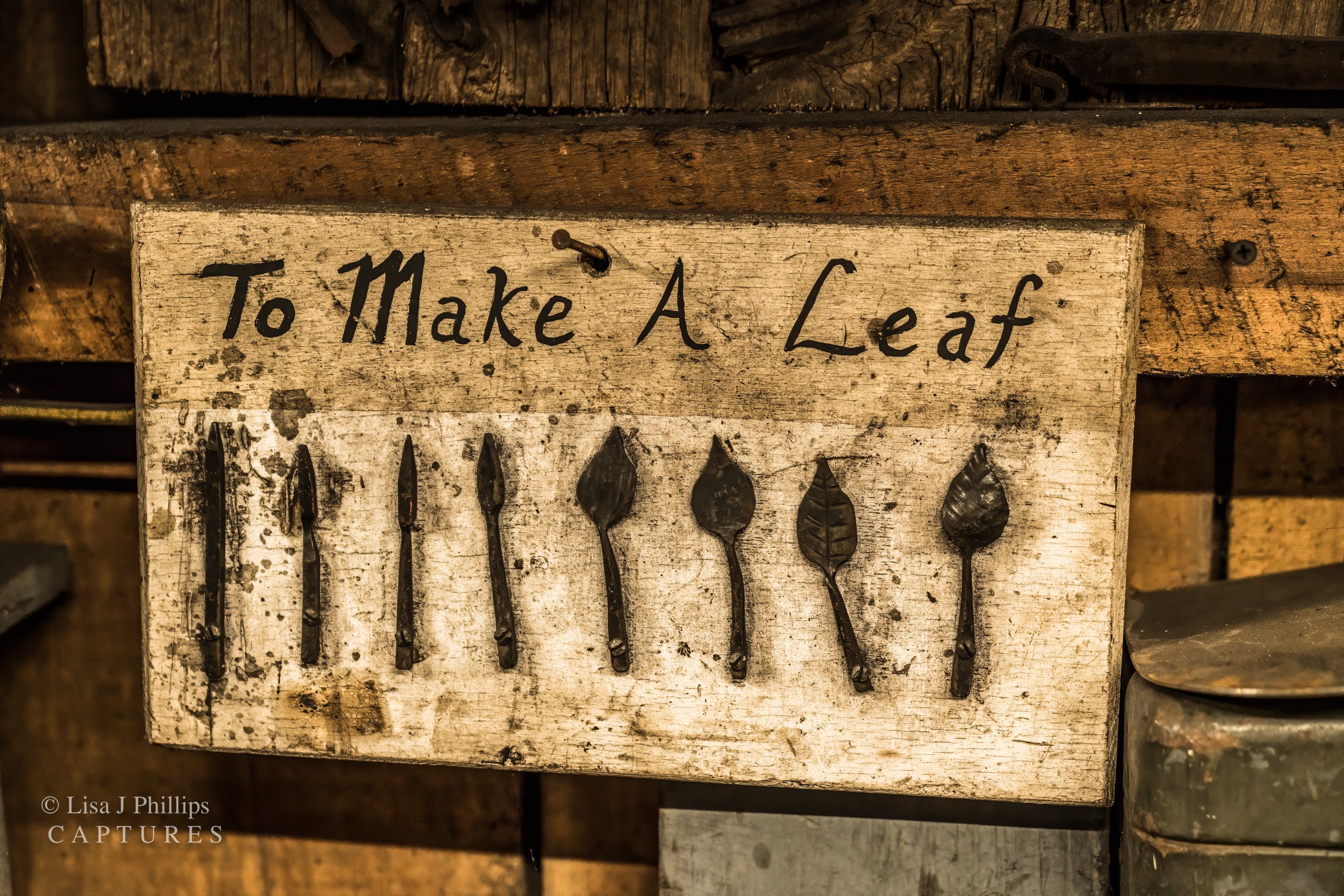 A wooden board nailed to a wall. It reads ‘To Make A Leaf’ across the top. Below are eight pieces of steel showing each of the steps to forging a leaf,